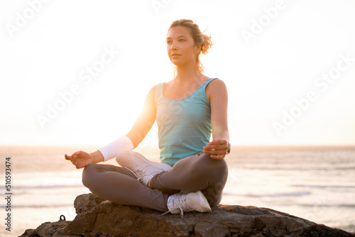 Woman practice yoga lotus pose to meditation with summer vacation rocky beach happiness and relaxation. Calm female exercise with yoga meditate ocean beach with sunset golden time.