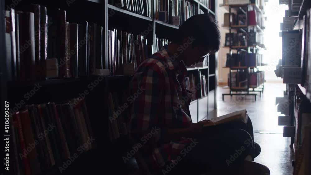 Male student on floor in the library in dim light reading book preparing for exams. Slide shot from behind shelf, at a guy sitting on the floor with a book Male student on floor in the library in dim light reading book preparing for exams. Slide shot from behind shelf, at a guy sitting on the floor with a book