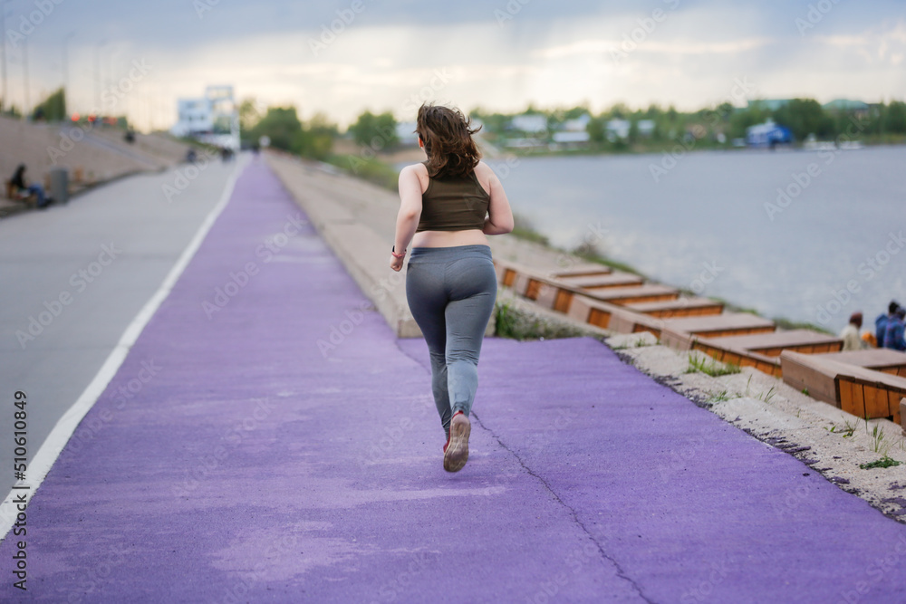 European teenage girl overweight on jog on treadmill along embankment of city, overweight and active lifestyle of teenager