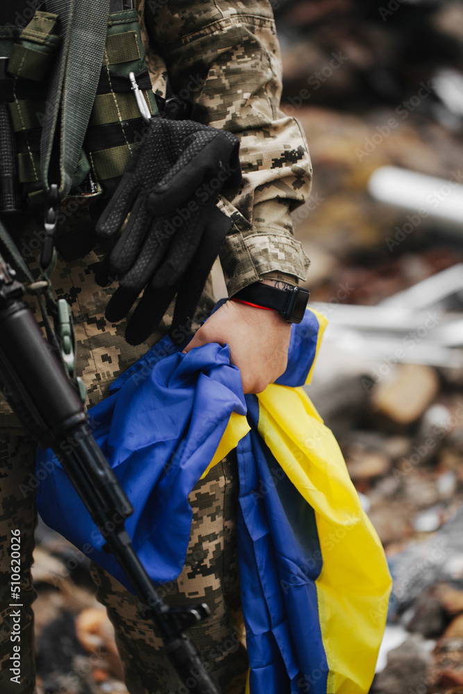Fototapeta premium Ukrainian military woman with the Ukrainian flag in her hands on the background of an exploded house