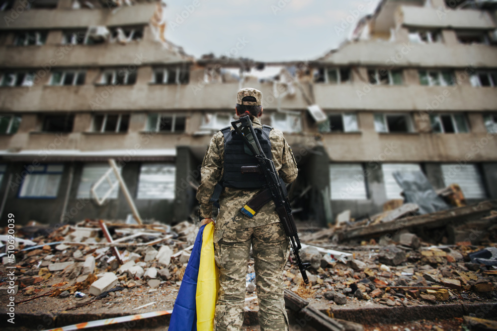 Ukrainian military woman with the Ukrainian flag in her hands on the background of an exploded house - obrazy, fototapety, plakaty 