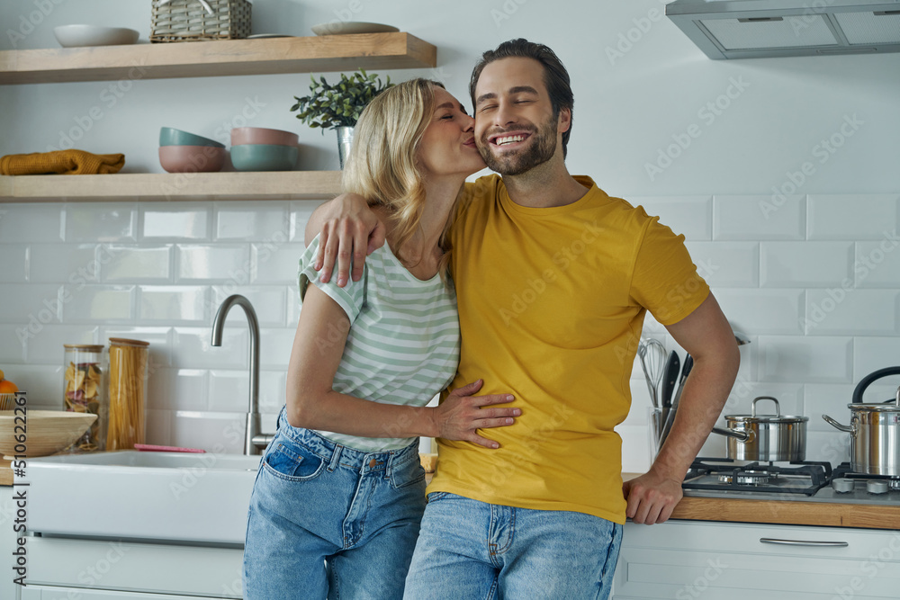 Cute couple embracing and kissing while standing at the domestic kitchen Cute couple embracing and kissing while standing at the domestic kitchen