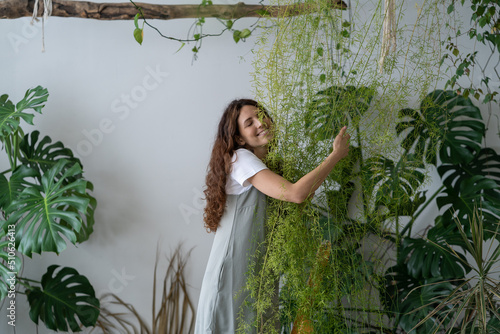 Obraz na plátně Young Spanish girl stands among green plants on stepladder and hugs asparagus in living room