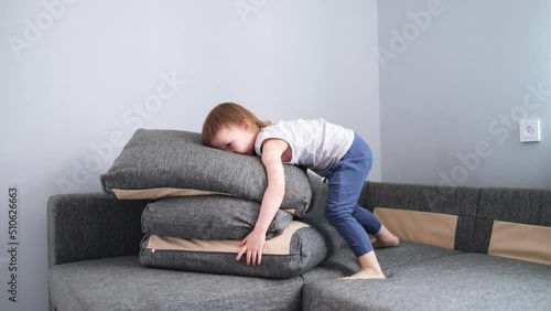 Funny toddler girl plays with pillows on couch, builds house and throws. Happy childhood, cozy interior
