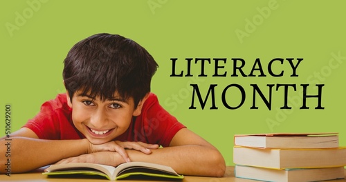 Portrait of smiling cute boy reading book on table and literacy month text on green background
