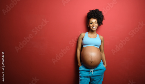 Portrait of blissful black pregnant woman on fitness sportswear against red background.
