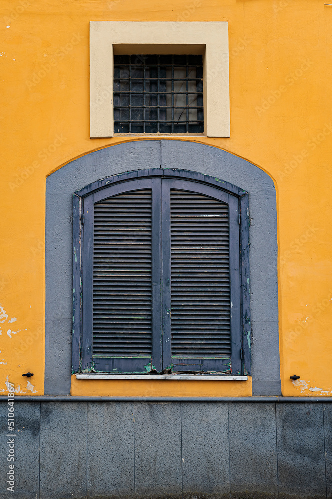 An old window with closed wooden shutters and one small wreath with a square lattice, like a prison on the yellow wall of the house
