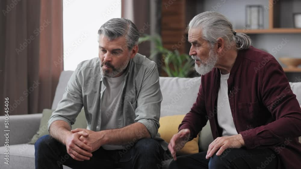 Family conflict. Adult son visiting senior father. Two generations family has unpleasant conversation sitting on couch at home. Generational confrontation. Family conflict. Adult son visiting senior father. Two generations family has unpleasant conversation sitting on couch at home. Generational confrontation.