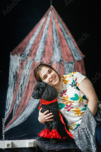 Black poodle dog in red outfit and collar with owner woman sits at painted tent, playing circus
