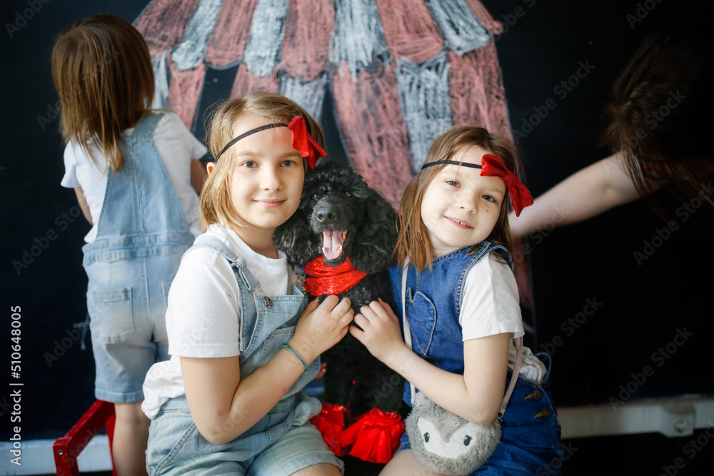 Fototapeta premium Children together with pet dog play in theater, circus. Black poodle and three European kids against background of black chalk board with circus pattern.