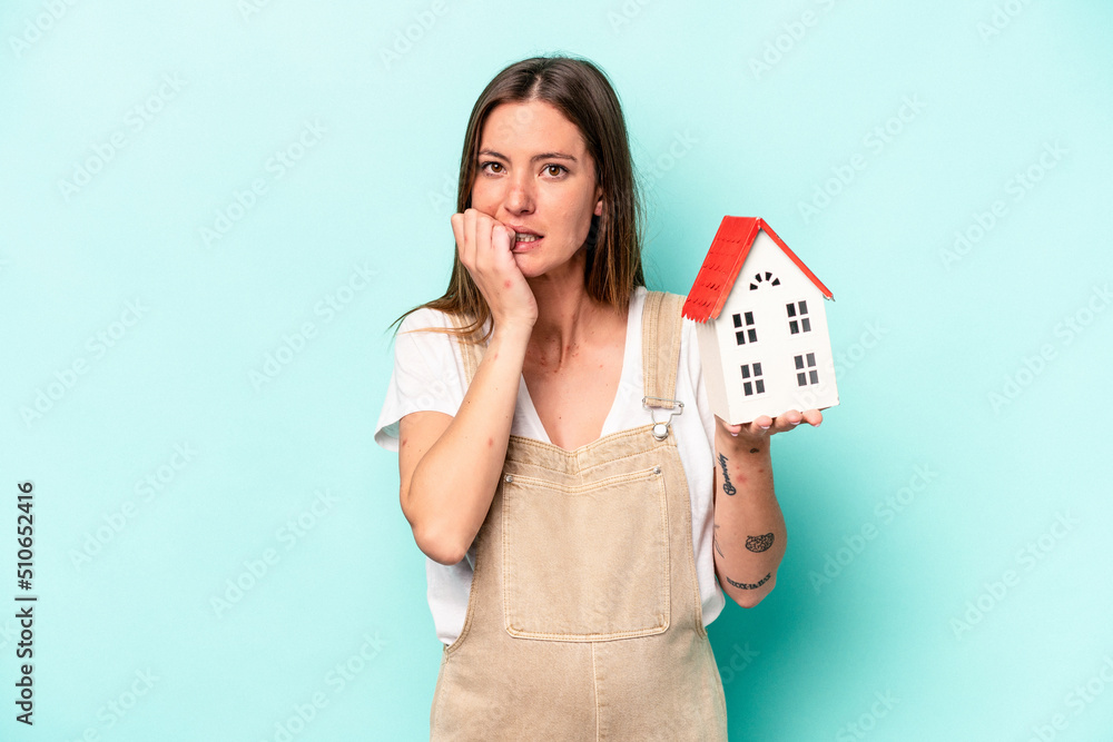 Young caucasian pregnant woman holding an toy house isolated on blue background biting fingernails, nervous and very anxious. Young caucasian pregnant woman holding an toy house isolated on blue background biting fingernails, nervous and very anxious.