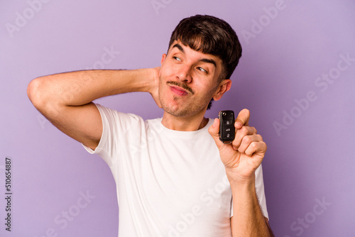 Young hispanic man holding keys car isolated on purple background touching back of head, thinking and making a choice.