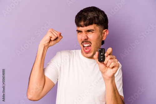 Young hispanic man holding keys car isolated on purple background raising fist after a victory, winner concept.