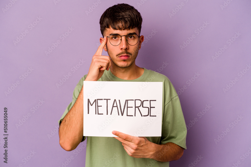 Young hispanic man holding meta verse placard isolated on purple background pointing temple with finger, thinking, focused on a task. Young hispanic man holding meta verse placard isolated on purple background pointing temple with finger, thinking, focused on a task.