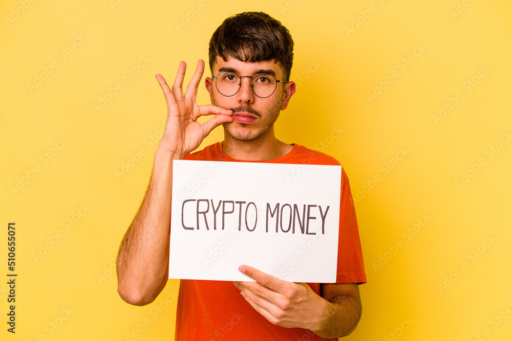 Young hispanic man holding nft placard isolated on yellow background with fingers on lips keeping a secret. Young hispanic man holding nft placard isolated on yellow background with fingers on lips keeping a secret.