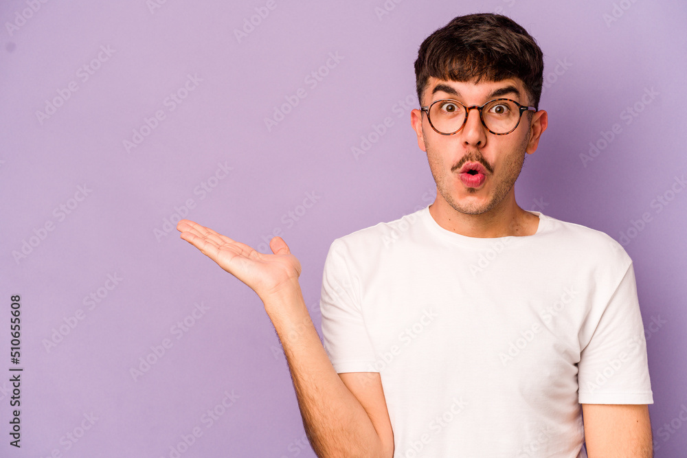 Fototapeta premium Young caucasian man isolated on purple background impressed holding copy space on palm.
