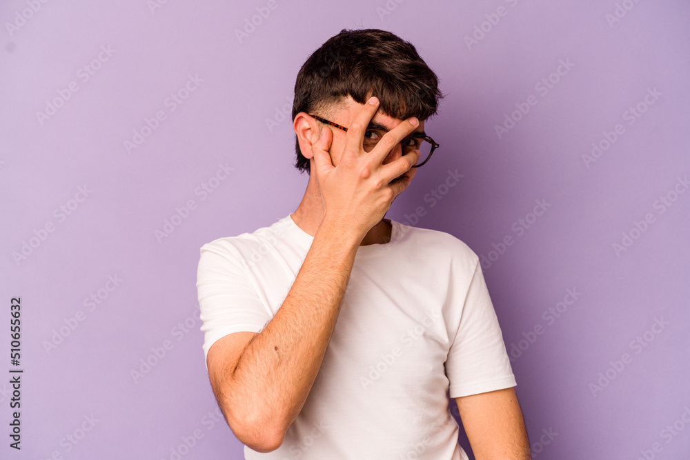 Young caucasian man isolated on purple background blink at the camera through fingers, embarrassed covering face. Young caucasian man isolated on purple background blink at the camera through fingers, embarrassed covering face.