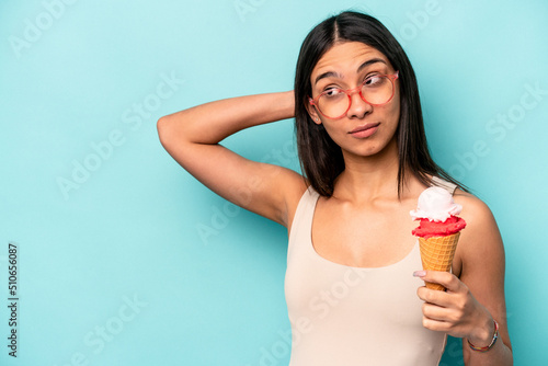 Young hispanic woman holding an ice cream isolated on blue background touching back of head, thinking and making a choice.