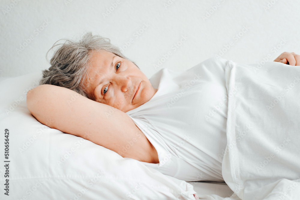 Side view of an awakened senior woman resting on bed in bedroom in morning. Elderly woman lying on pillow and looking at camera Side view of an awakened senior woman resting on bed in bedroom in morning. Elderly woman lying on pillow and looking at camera