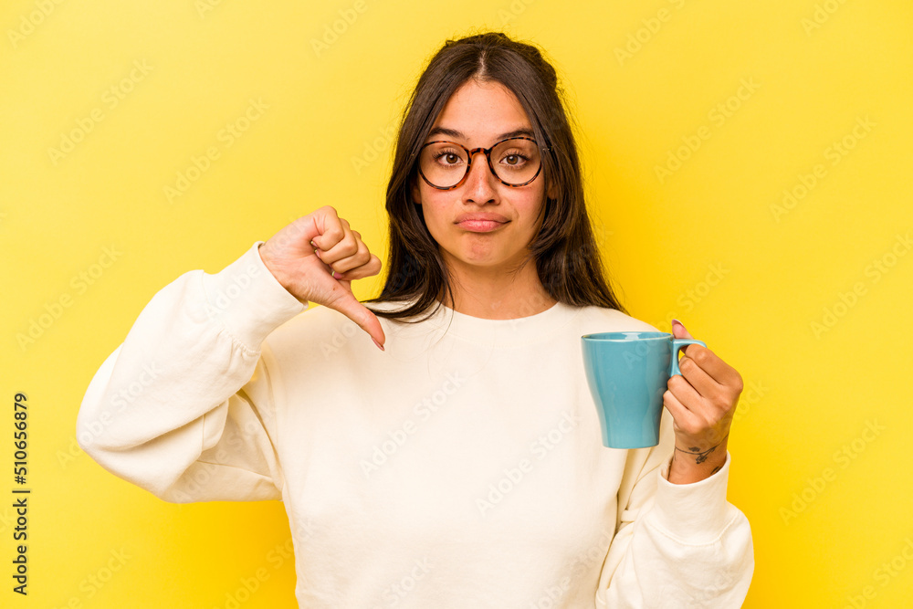 Young hispanic woman holding a mug isolated on yellow background showing a dislike gesture, thumbs down. Disagreement concept. Young hispanic woman holding a mug isolated on yellow background showing a dislike gesture, thumbs down. Disagreement concept.