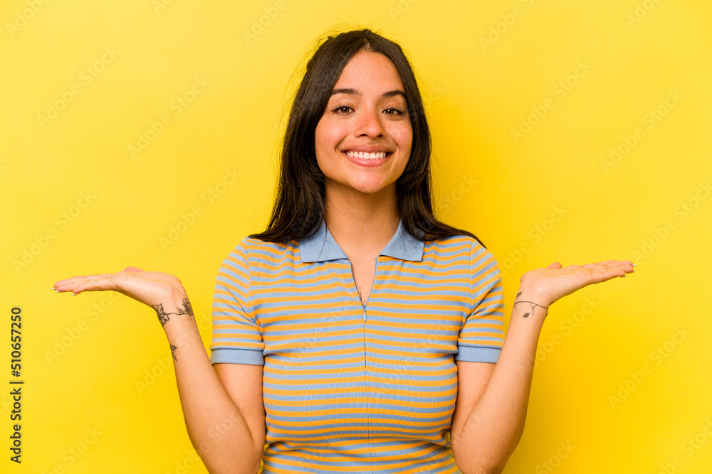 Young hispanic woman isolated on yellow background makes scale with arms, feels happy and confident. Young hispanic woman isolated on yellow background makes scale with arms, feels happy and confident.