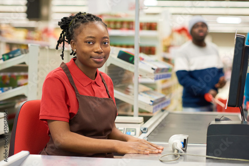 Happy young black woman in red shirt and brown apron sitting by counter with scanner and computer screen showing price of goods