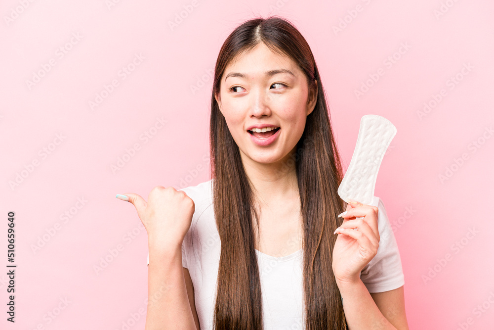 Young asian woman holding sanitary napkin isolated on pink background points with thumb finger away, laughing and carefree. Young asian woman holding sanitary napkin isolated on pink background points with thumb finger away, laughing and carefree.