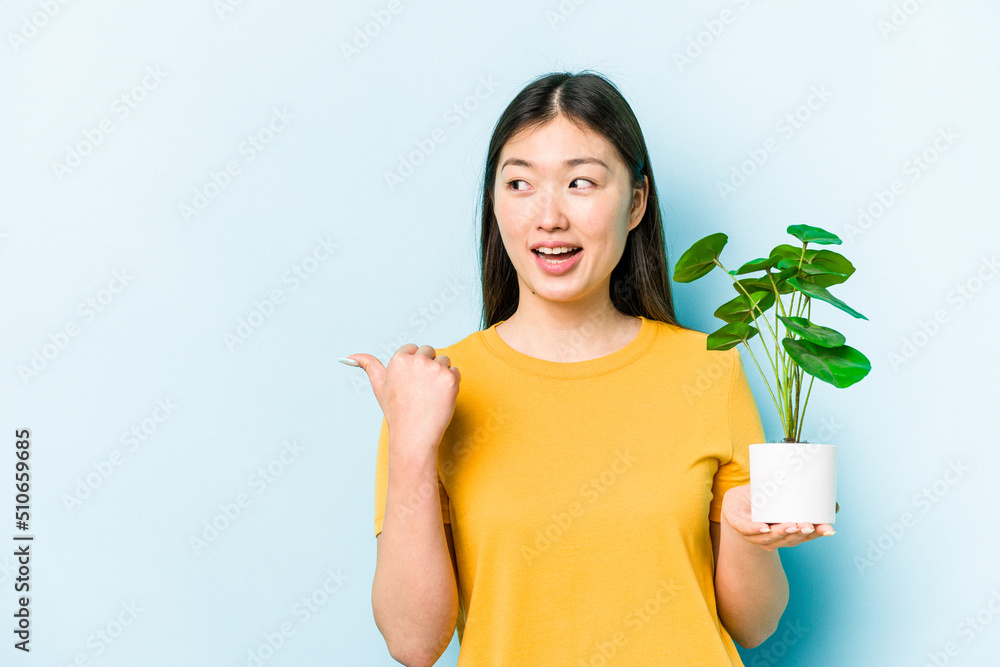 Young asian woman holding a plant isolated on blue background points with thumb finger away, laughing and carefree.