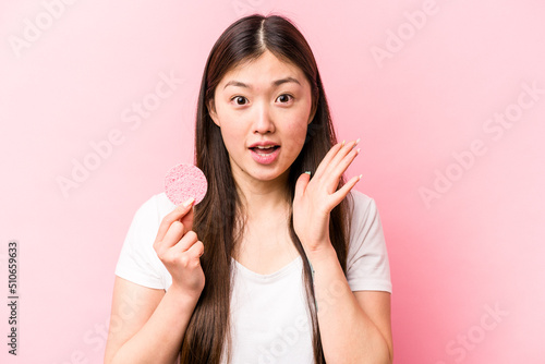 Young asian woman holding facial sponge isolated on pink background surprised and shocked.