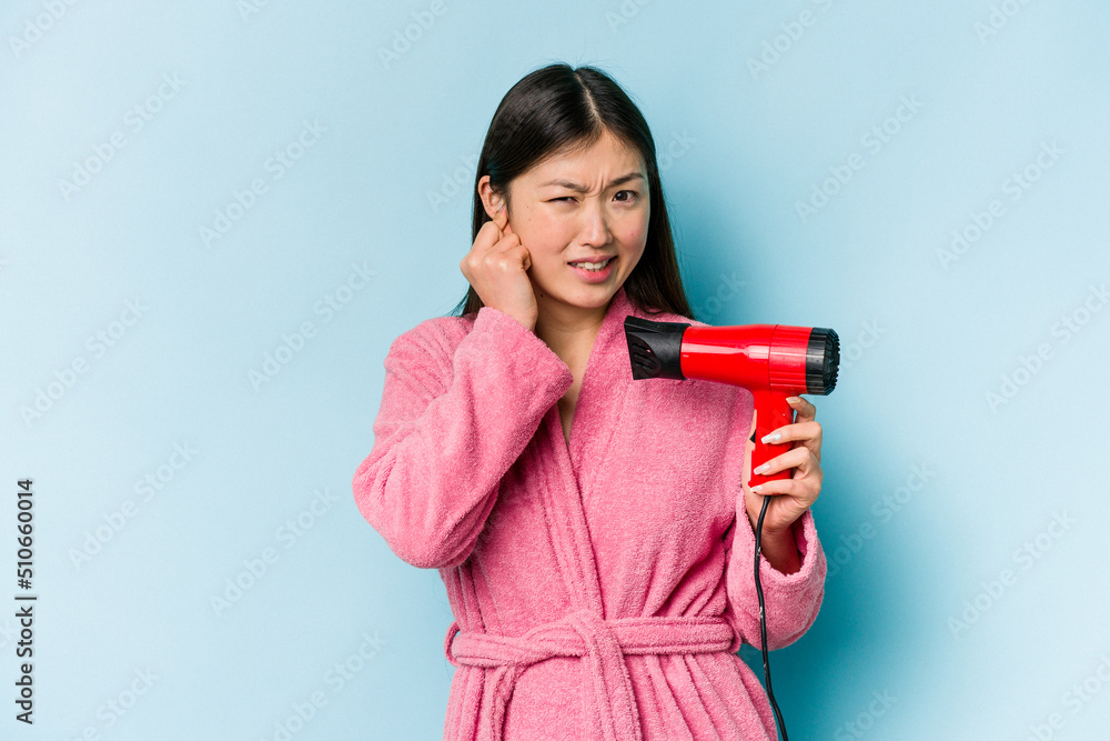 Young asian woman wearing a bathrobe and holding hairdryer isolated on pink background covering ears with hands. Young asian woman wearing a bathrobe and holding hairdryer isolated on pink background covering ears with hands.