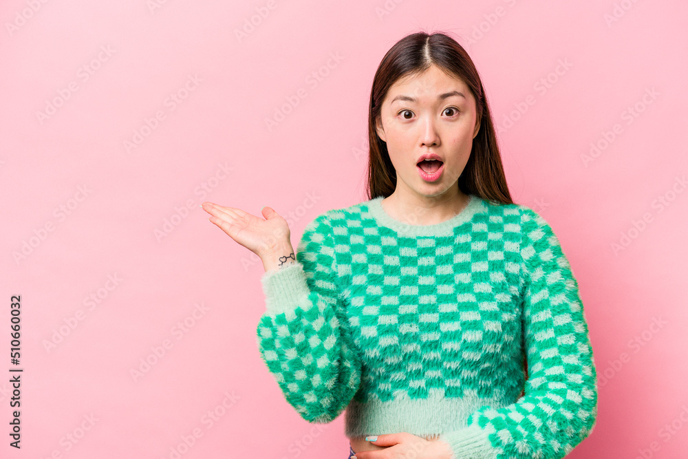 Young Chinese woman isolated on pink background impressed holding copy space on palm. Young Chinese woman isolated on pink background impressed holding copy space on palm.