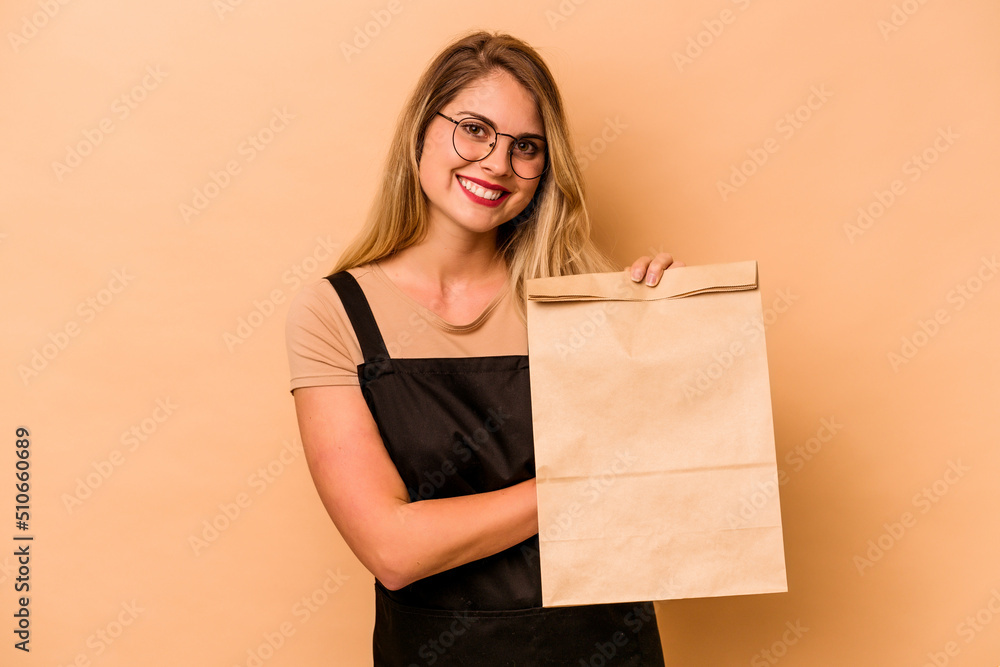 Restaurant waiter caucasian woman holding a take away bag isolated on beige background laughing and having fun. Restaurant waiter caucasian woman holding a take away bag isolated on beige background laughing and having fun.
