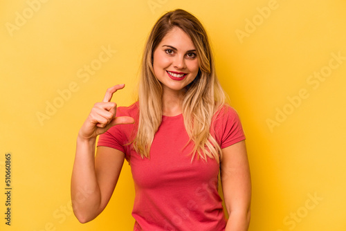 Young caucasian woman isolated on yellow background holding something little with forefingers, smiling and confident.