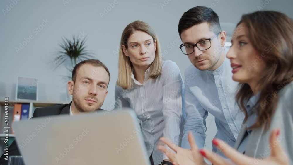 Group of young Caucasian colleagues sitting at silver laptop and looking at screen. Beautiful Ukrainian woman in foreground explaining concept of presentation. Coworkers in formal clothes listening Group of young Caucasian colleagues sitting at silver laptop and looking at screen. Beautiful Ukrainian woman in foreground explaining concept of presentation. Coworkers in formal clothes listening