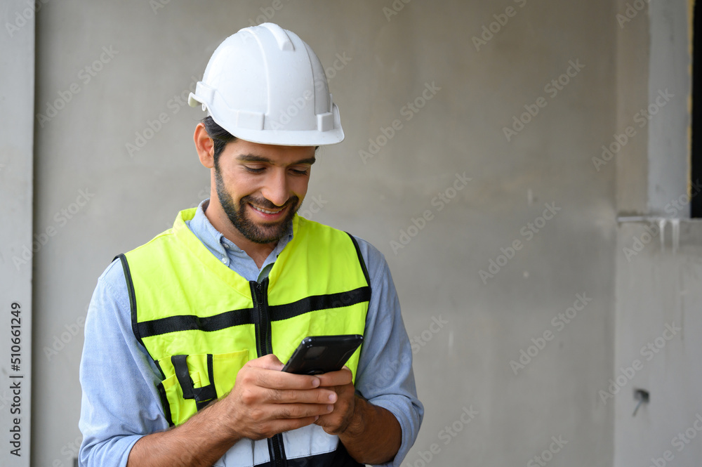 portrait of young engineer in vest with white helmet standing on construction site, smiling and holding smartphone for worker, internet, social media. portrait of young engineer in vest with white helmet standing on construction site, smiling and holding smartphone for worker, internet, social media.