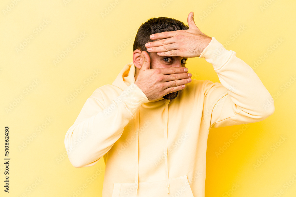 Young caucasian man isolated on yellow background blink at the camera through fingers, embarrassed covering face. Young caucasian man isolated on yellow background blink at the camera through fingers, embarrassed covering face.