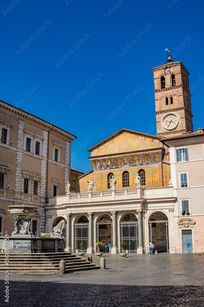 The basilica of Santa Maria in Trastevere, in a square of the  center of Rome. The facade with the portico and the mosaic, the bell tower with the clock and the fountain with the inscription SPQR.