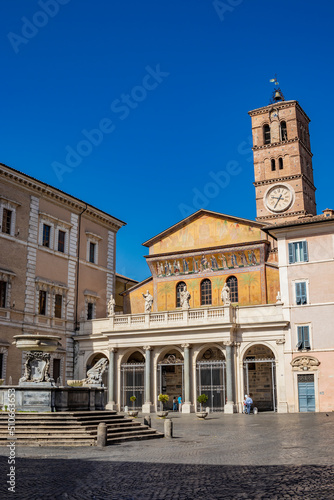 The basilica of Santa Maria in Trastevere, in a square of the center of Rome. The facade with the portico and the mosaic, the bell tower with the clock and the fountain with the inscription SPQR.