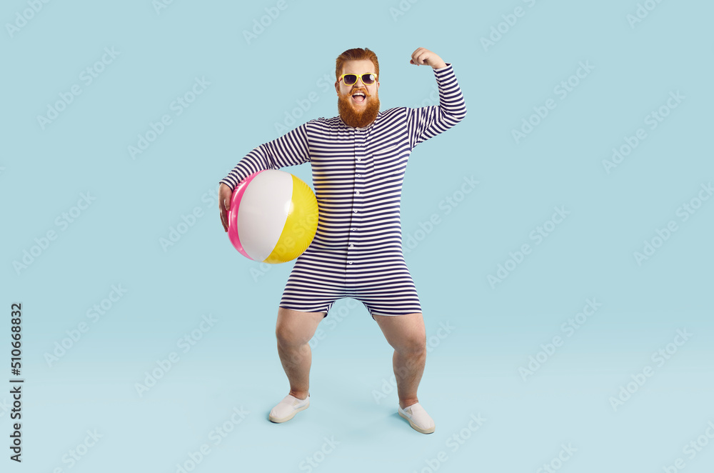 Cheerful funny chubby man with inflatable swimming ball shows muscles on light blue background. Full length portrait of humorous strong bearded man in striped leotard and sunglasses. Banner.