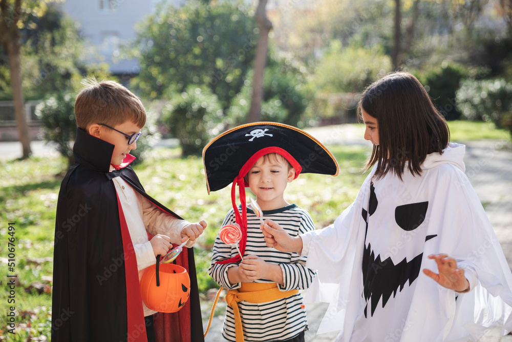 Treat or trick! Happy kids wearing carnival costumes of ghost, pirate and vampire for Halloween celebration sharing candies with each other staying in the park.