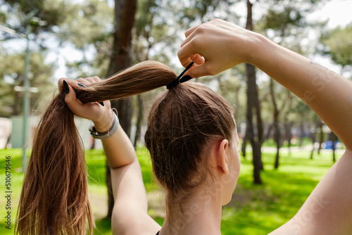 Beautiful brunette woman wearing sports bra standing on city park, outdoors tying her hair in a ponytail while looking away.