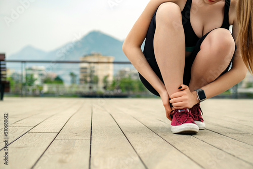 Brunette sportive woman wearing black sports bra standing on city park  outdoors suffering from an ankle injury while exercising and running. Healthcare and sport concept. Ankle sprained.