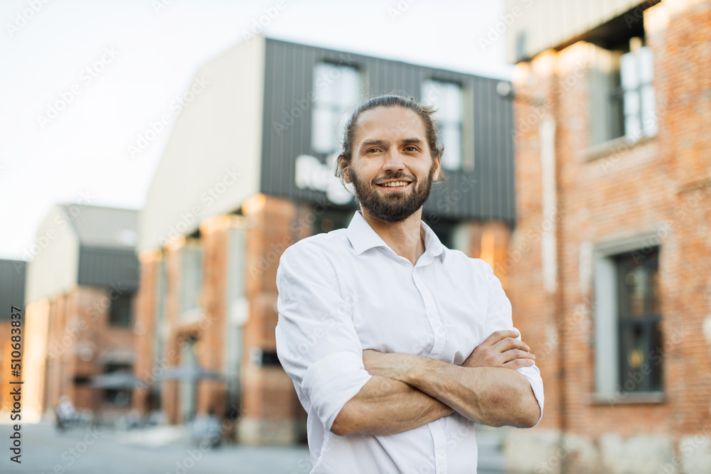 Portrait of young handsome smiling bearded man with crossed arms on background of city. Charming Caucasian businessman standing near office space during break. Portrait of young handsome smiling bearded man with crossed arms on background of city. Charming Caucasian businessman standing near office space during break.