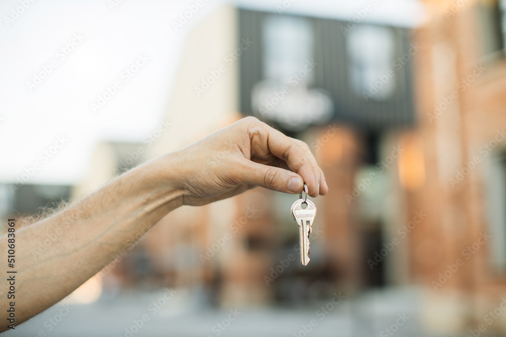 House keys in male hand on background of new buildings and sky. Real estate agent, moving home or renting property. Man hand holds the keys to an apartment against the background of buildings.