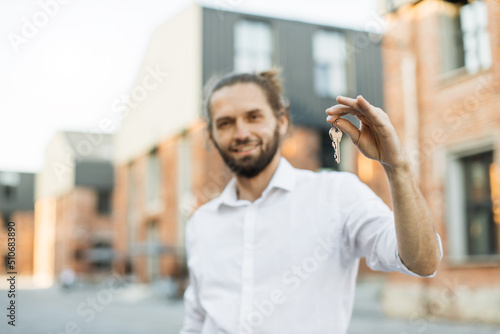 Focus on hand with keys. Handsome young businessman in white shirt holding keys of new flat, looking at camera and smiling, standing near the building in the middle of the street.