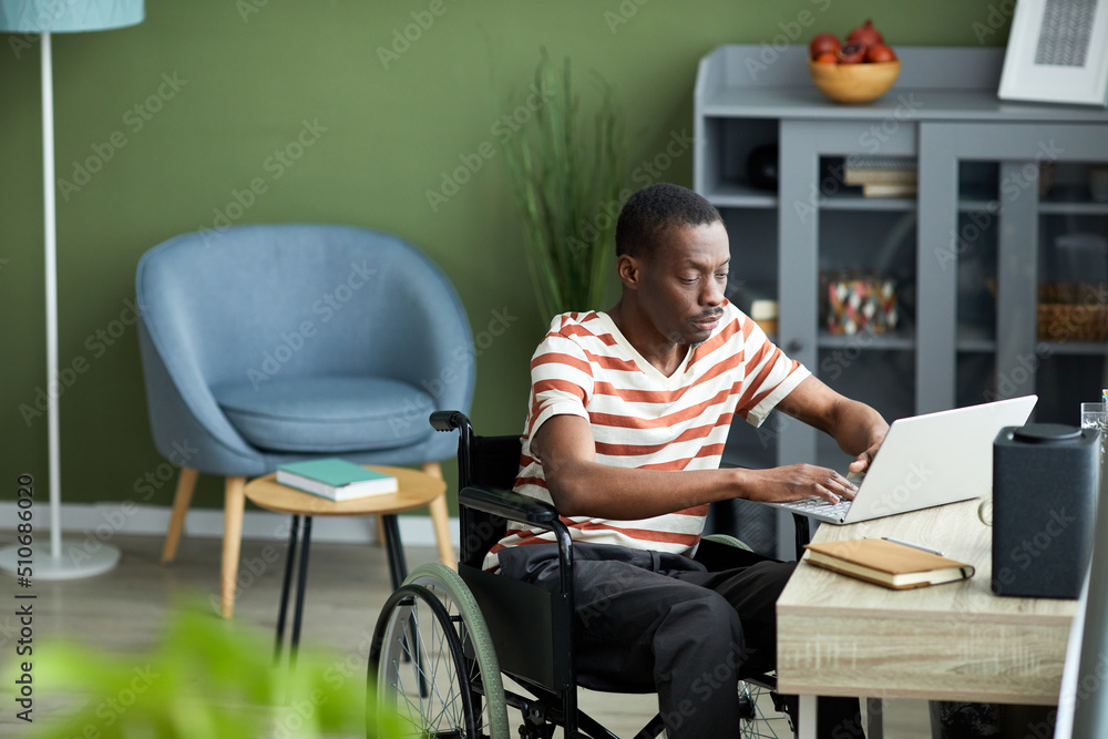 Portrait of African American man with disability using laptop while working from home against green wall, copy space