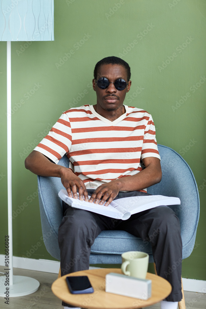 Vertical portrait of adult black man reading tactile book in braille while relaxing at home in armchair Vertical portrait of adult black man reading tactile book in braille while relaxing at home in armchair