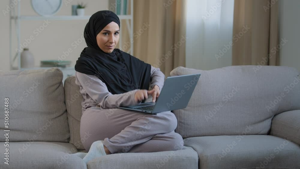 Attractive young muslim woman wear hijab sit on cozy sofa in living room adorable energetic millennial islamic arabian girl has fun in living room move hands beat rhythm on laptop enjoy rest free time Attractive young muslim woman wear hijab sit on cozy sofa in living room adorable energetic millennial islamic arabian girl has fun in living room move hands beat rhythm on laptop enjoy rest free time