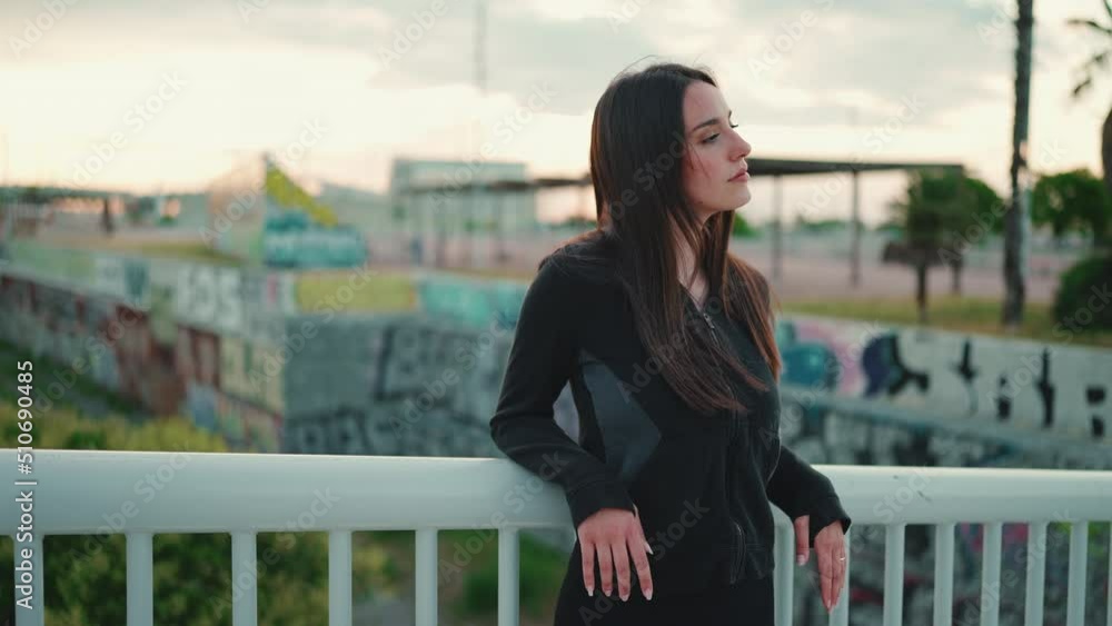 Pretty woman with freckles and loose brown hair wearing black sports hoodie stands on bridge and looks away turns her head to the camera and smiles. Сamera moving Pretty woman with freckles and loose brown hair wearing black sports hoodie stands on bridge and looks away turns her head to the camera and smiles. Сamera moving
