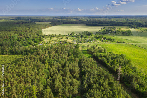 Pola i łąki wiosną widziane z dużej wysokości. Zdjęcie z drona. Rozległy, płaski teren pokryty zielonymi polami uprawnymi i łąkami. Widać polną drogę, kępy drzew, oraz na obrzeżach iglasty las. Widok